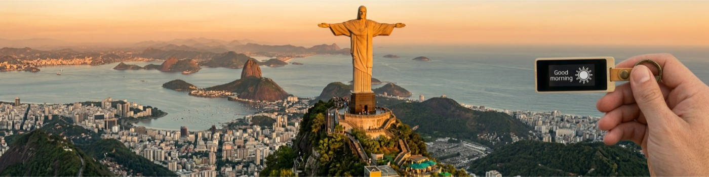Christ the Redeemer overlooking Rio de Janeiro at sunset, with a KEEP bamboo keychain held in the foreground showing a 'Good morning' note on its e-ink screen.