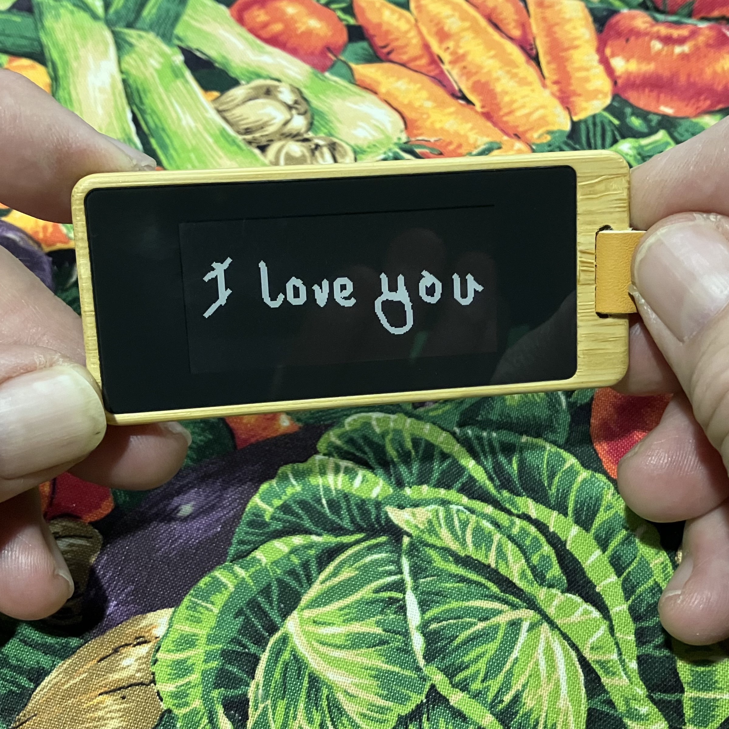 A beta tester's hands holding the black-and-white KEEP keychain, the e-ink screen showing a handwritten 'I love you'.